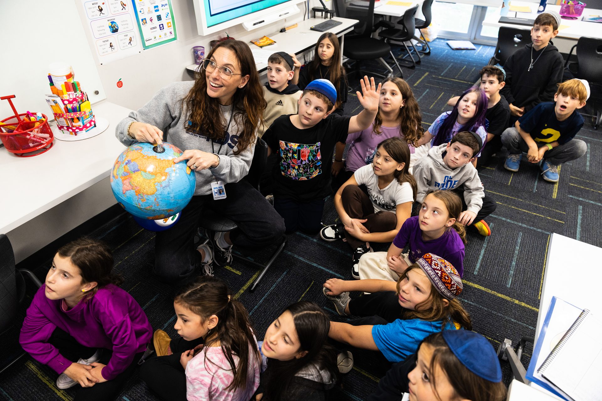 A Hillel Day School teacher leading a lesson with a globe as students raise their hands